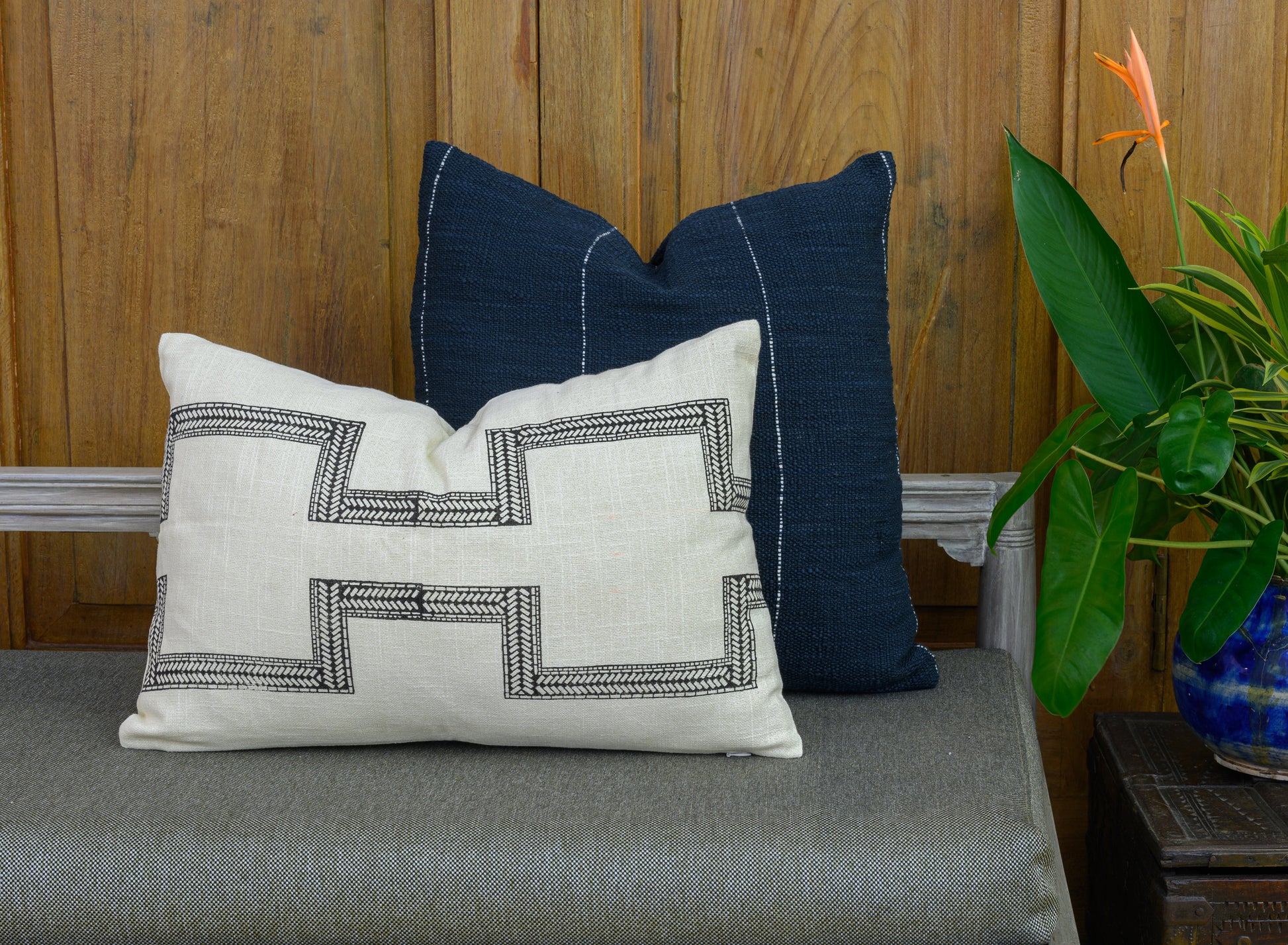 Mood shot of a hand blocked cotton lumbar cushion cover in a charcoal colour on an ivory cotton fabric, placed in  front of a hand woven navy cushion cover on a bench